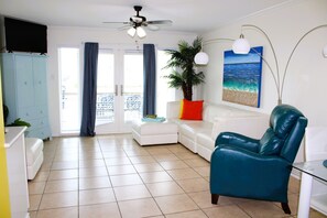 Ein Wohnzimmer mit einem weißen Sofa, einem blaugrünen Sessel, einem Deckenventilator, einem Topf mit Pflanze und einem Gemälde vom Meer.