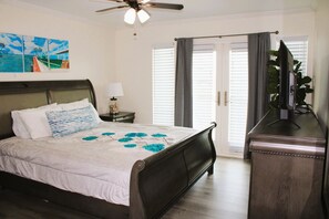 Ein Schlafzimmer mit einem Bett, Deckenventilator, Nachttisch, Kommode, Fernseher und einem Fenster mit Vorhängen.