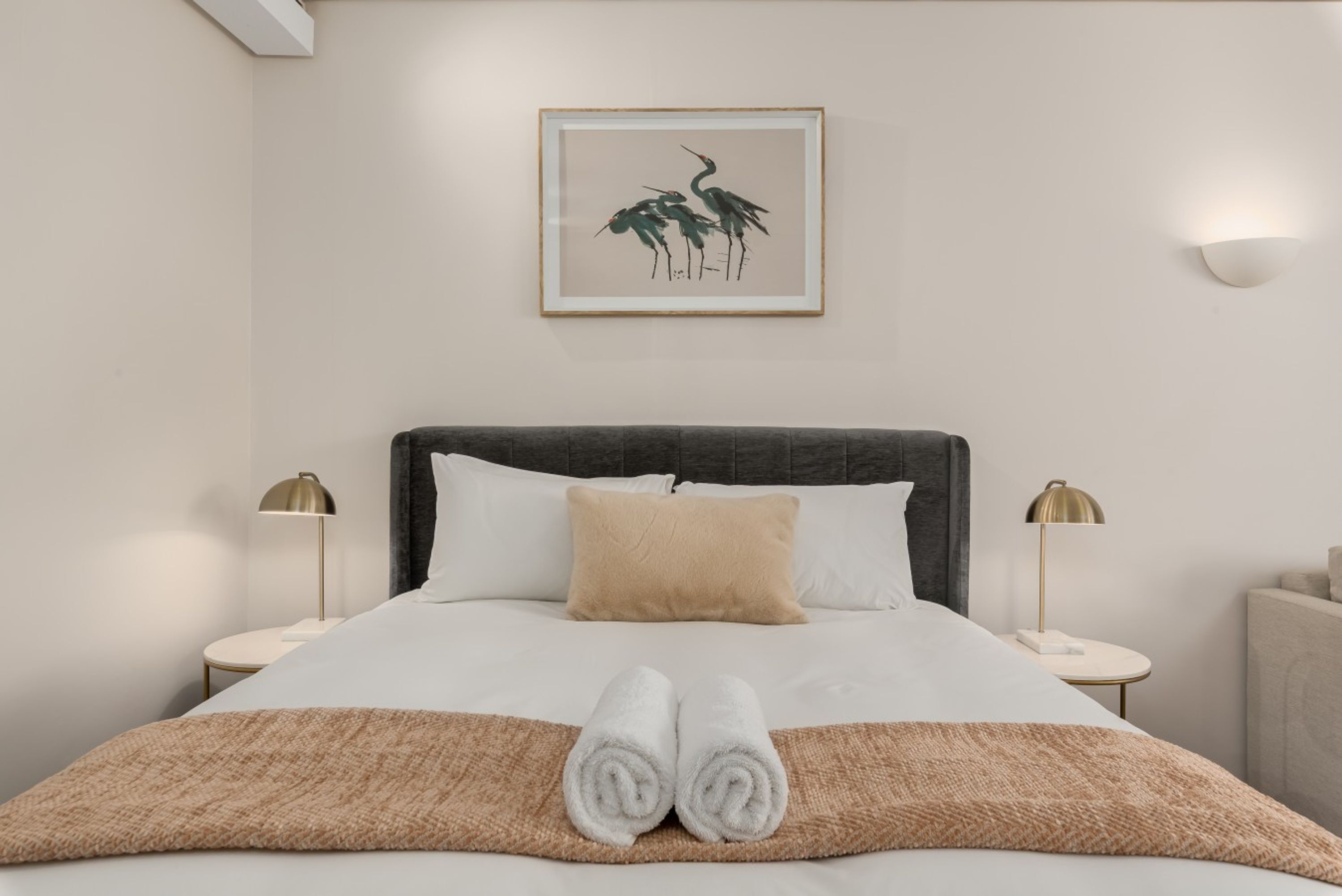 Un lit bien fait, recouvert d’un plaid marron, avec deux lampes de chevet et une œuvre d’art encadrée accrochée au mur.