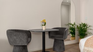 A modern room with a round table, two grey chairs, a small potted plant with yellow flowers, a large mirror, and a bed with a white blanket. - Bella at Dolce Vita | Stylish Sea Point Studio (Cape Town)