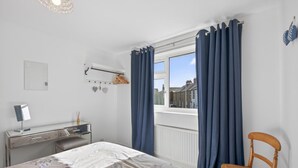 Ett sovrum med en säng, ett skrivbord, en stol, ett fönster med gardiner, ett element och utsikt över byggnader utanför.