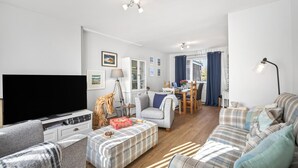 Ett modernt vardagsrum med en platt-TV, en soffa, ett matbord med stolar och ett köksområde i bakgrunden.