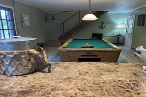 A pool table in a room with a staircase, a hanging light fixture, and a decorative anchor on the wall. - Wight Tide Landing (Midway)
