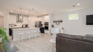 Una cocina moderna con armarios blancos, una isla central con taburetes, un sofá oscuro y un televisor montado en la pared.