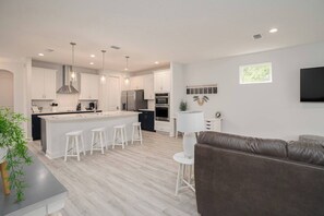 A modern kitchen with white cabinets, a central island with stools, a dark sofa, and a TV mounted on the wall. - Close to Vilano Beach - Private Hot Tub (St. Augustine)