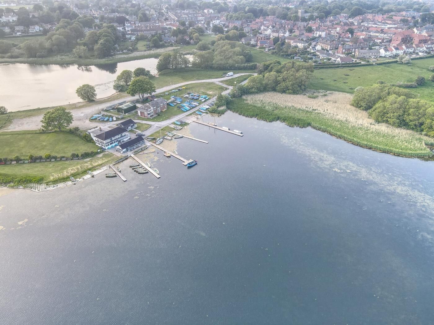 Vue aérienne d’une propriété au bord du lac, avec un quai, des bateaux et un bâtiment doté d’une grande fenêtre.