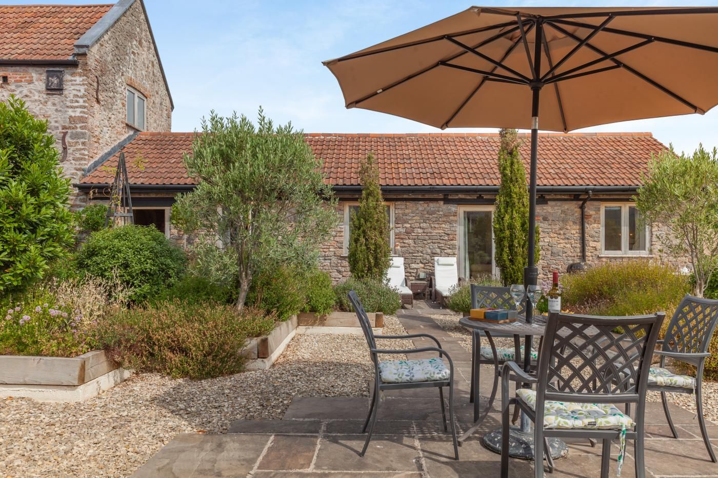 A patio area with outdoor furniture and a large umbrella, surrounded by stone buildings and greenery.