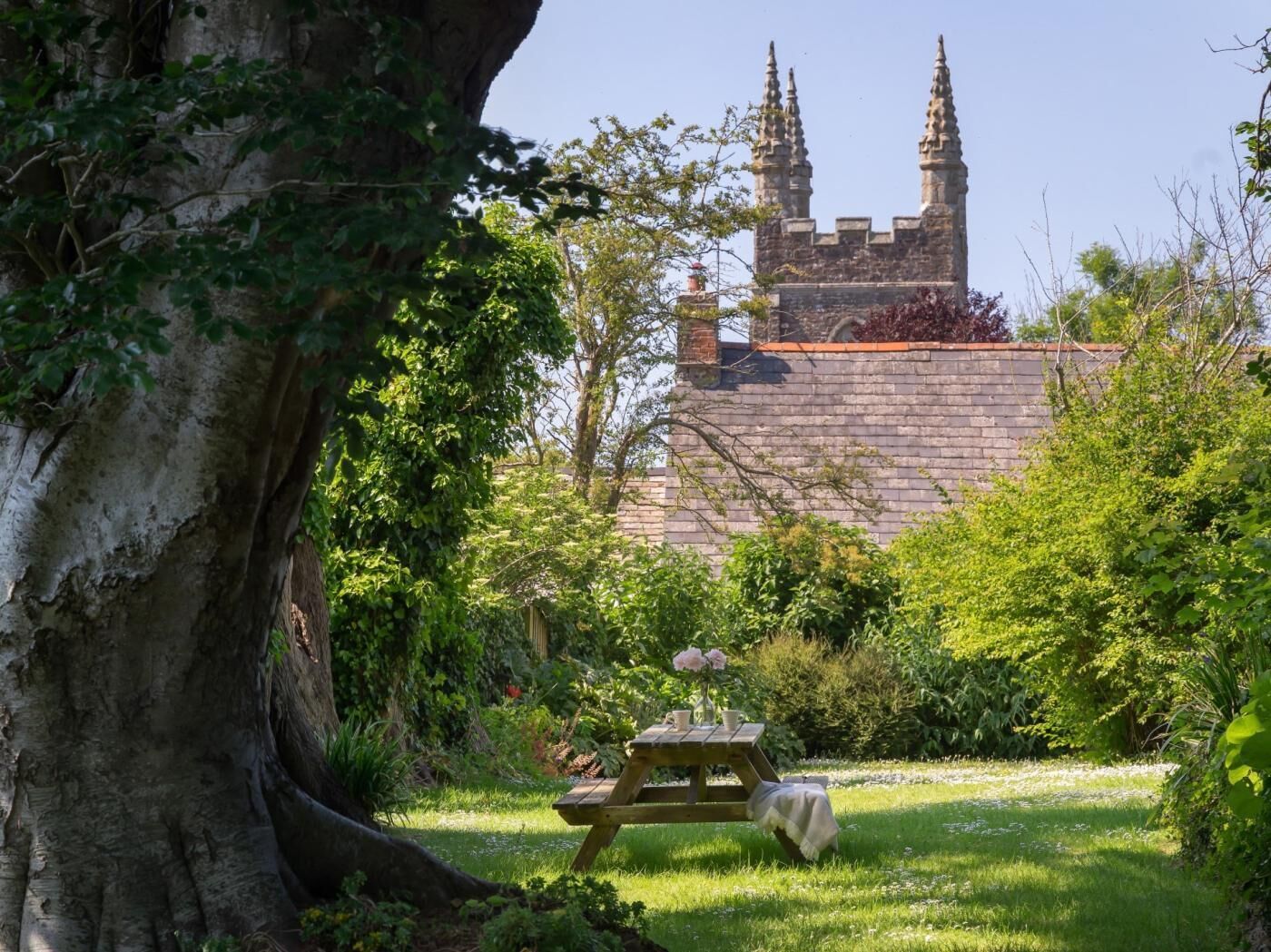 A stone building with two towers, surrounded by lush greenery and a picnic table with flowers.