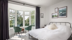 Quarto com uma cama grande, uma escrivaninha, duas cadeiras e vista para o verde através de janelas amplas.
