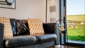 A modern living room with a leather sofa, patterned cushions, a floor lamp, and a view of a green field through a large window. - 2 Bed in Bude (oc-86152) (Widemouth Bay)