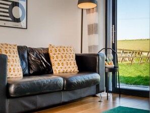 Ein modernes Wohnzimmer mit einem Ledersofa, gemusterten Kissen, einer Stehlampe und Blick auf ein grünes Feld durch ein großes Fenster.