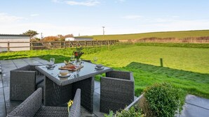 An outdoor patio with a table set for dining, surrounded by wicker chairs, overlooking a grassy field. - 2 Bed in Bude (oc-86152) (Widemouth Bay)