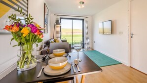 A dining area with a table set for two, a vase of flowers, and a view of the outdoors. - 2 Bed in Bude (oc-86152) (Widemouth Bay)