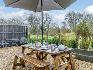 Een houten tuintafel gedekt met borden, bekers en een theepot, beschut onder een grote parasol, met een tuin en een houten hek op de achtergrond.
