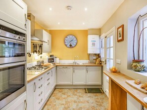 A kitchen with white cabinets, a clock, a stove, and a window with a plant. - 3 Bed in Keswick (oc-86162) (Keswick)