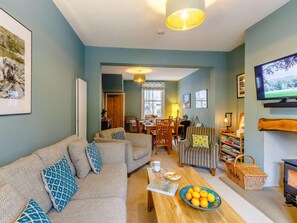 A cozy living room with a sofa, armchairs, a coffee table with oranges, a TV, and a fireplace. - 3 Bed in Keswick (oc-86162) (Keswick)