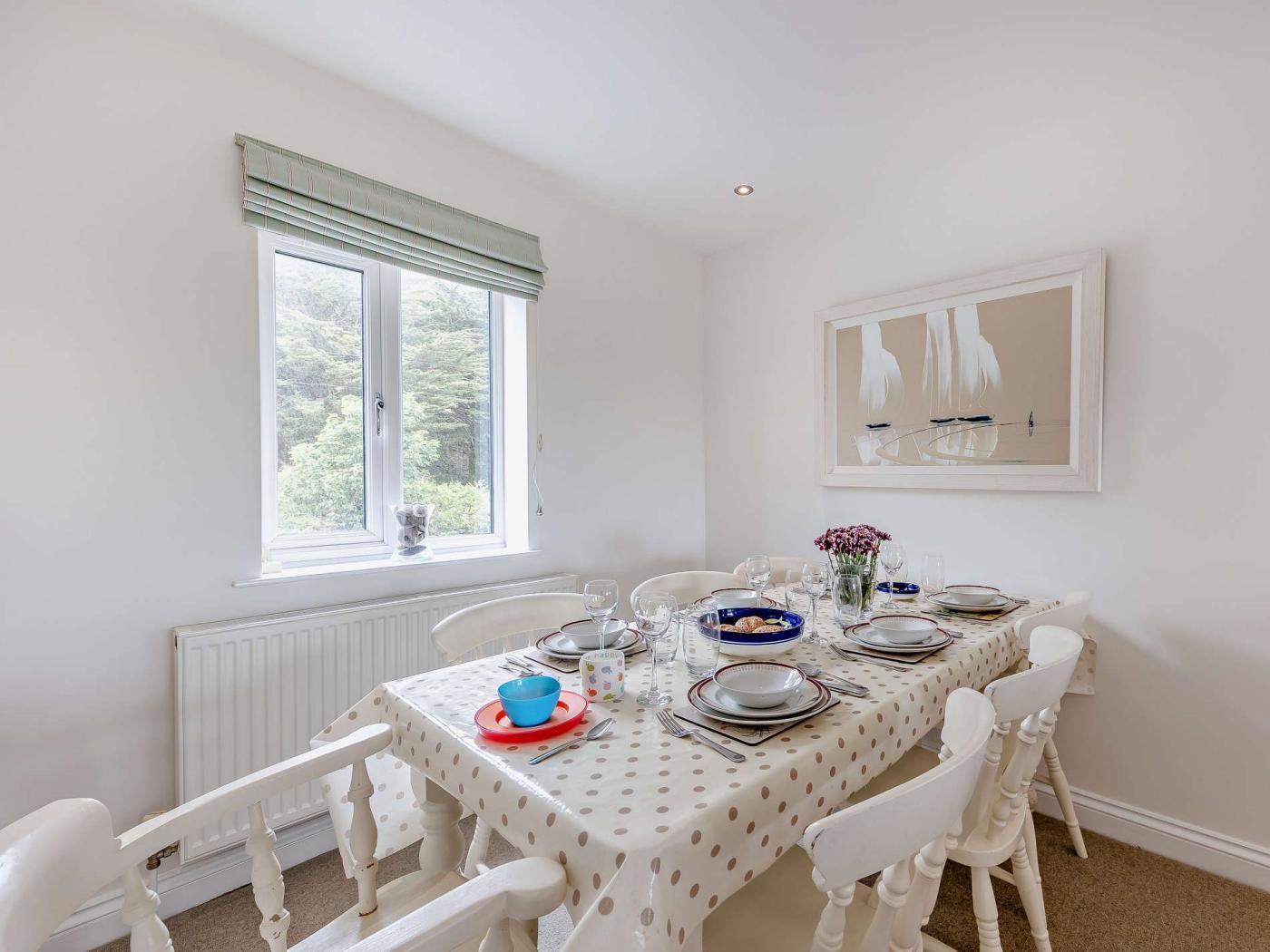 Een eetkamer met een tafel gedekt voor zes personen, witte stoelen, een raam met jaloezieën en een kunstwerk in een lijst aan de muur.