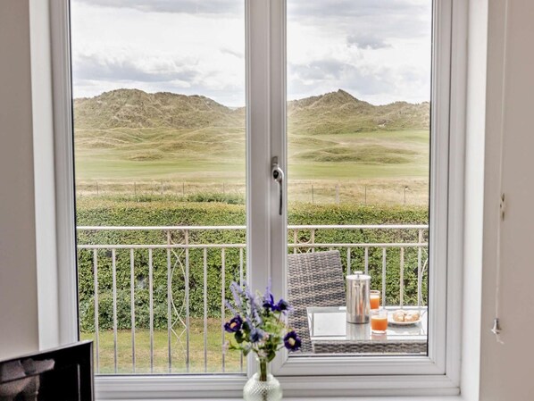 A room with a view of a grassy field and hills. There is a table with a vase of flowers and a metal container. - 3 Bed in Aberdyfi (oc-dy007) (Aberdovey)