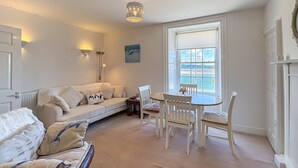 Ein gemütliches Wohnzimmer mit einer Couch, einem Esstisch mit Stühlen, einer Stehlampe und Meerblick durch ein großes Fenster.