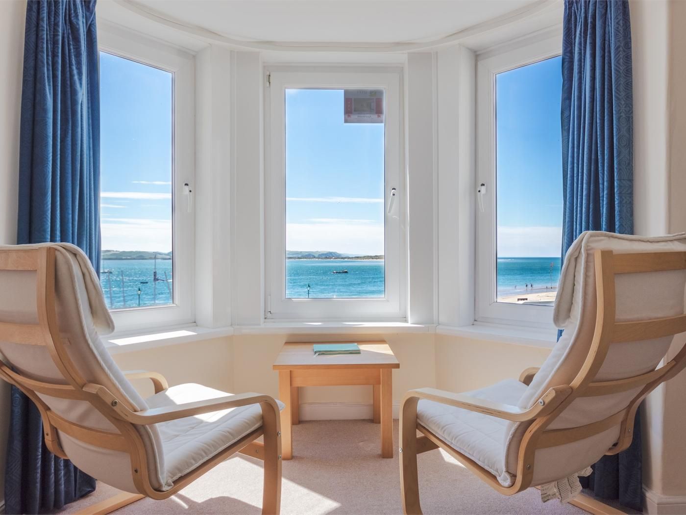 A room with a bay window overlooking a beach, two wooden chairs, a small table, and a view of the sea.