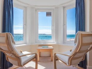 Ein Zimmer mit Erkerfenster und Blick auf einen Strand, zwei Holzstühle, ein kleiner Tisch und Meerblick.
