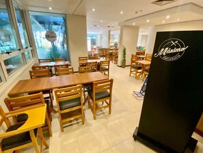A restaurant interior with wooden tables and chairs, a black sign with 'Maximo Restaurante' logo, and a view through the window.