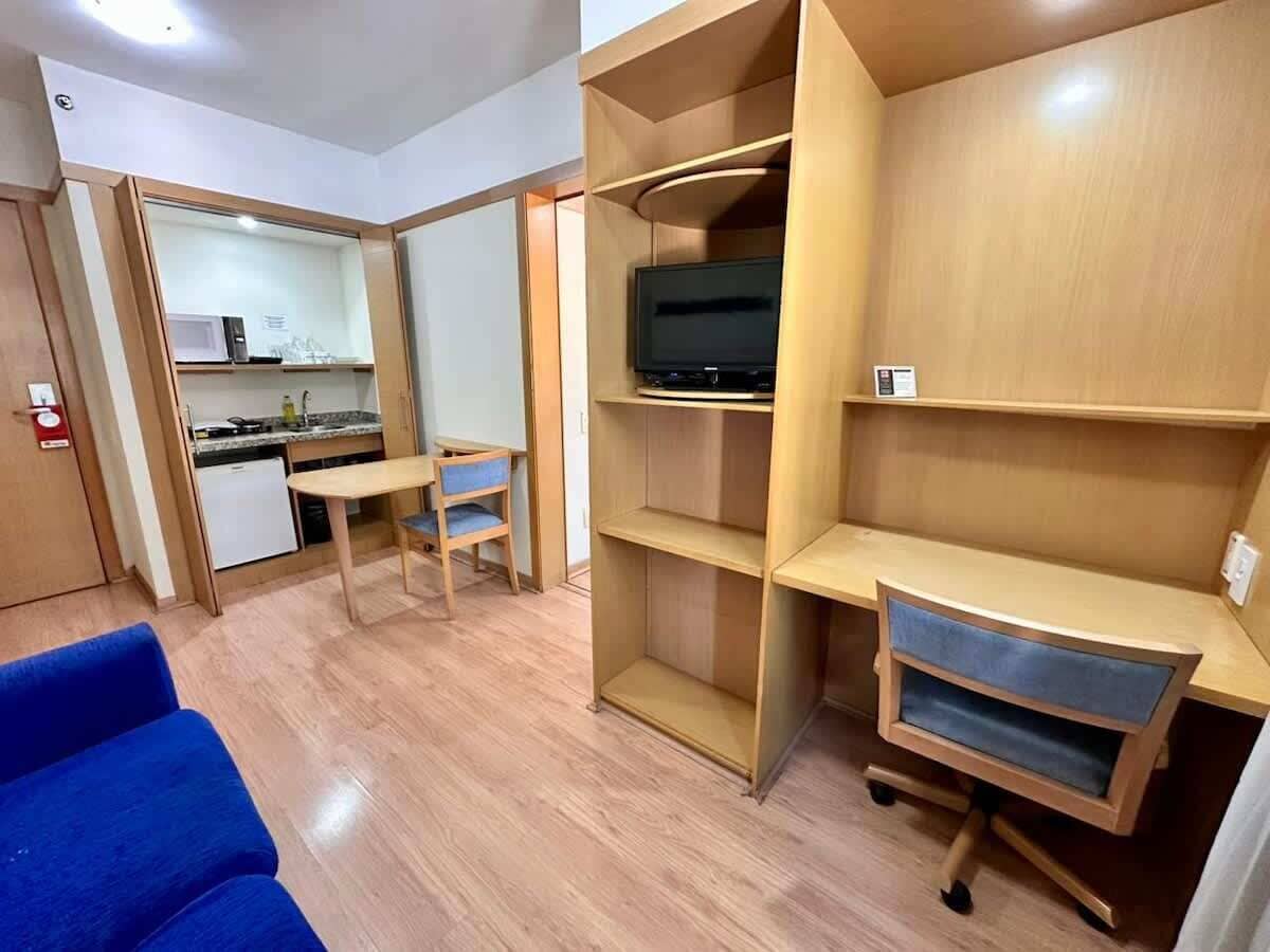 A compact room with a kitchenette, a television on a shelf, a blue sofa, and a desk with drawers.