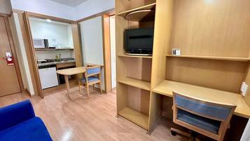A compact room with a kitchenette, a television on a shelf, a blue sofa, and a desk with drawers.