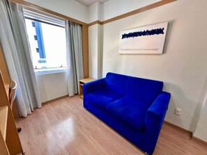 A room with a blue sofa, a window with curtains, a wall-mounted artwork, and a wooden floor.
