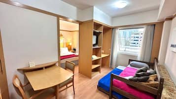A compact room with a bed, a desk, and a chair. There is a window with curtains, and a shelf unit is visible.