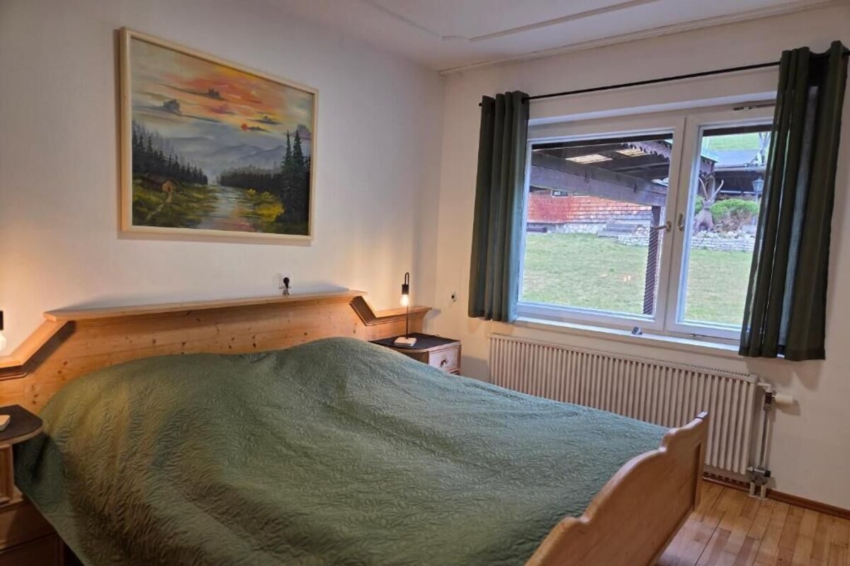 Ein Schlafzimmer mit einem hölzernen Bett, einem Nachttisch, einem Bild an der Wand und einem Fenster mit Vorhängen.