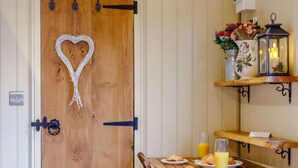Une porte en bois ornée d’un motif en forme de cœur, une table de petit-déjeuner avec des croissants et du jus d’orange, et une étagère avec des fleurs et une lanterne.