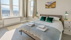 Ein Schlafzimmer mit einem großen Bett, zwei Nachttischen, Blick durch die Fenster und einem Bild an der Wand.