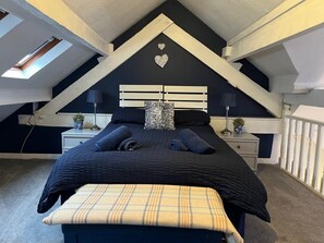 Ein Schlafzimmer mit schrägem Dach, einem Bett mit dunkler Bettwäsche, einer herzförmigen Dekoration an der Wand und einem Dachfenster.