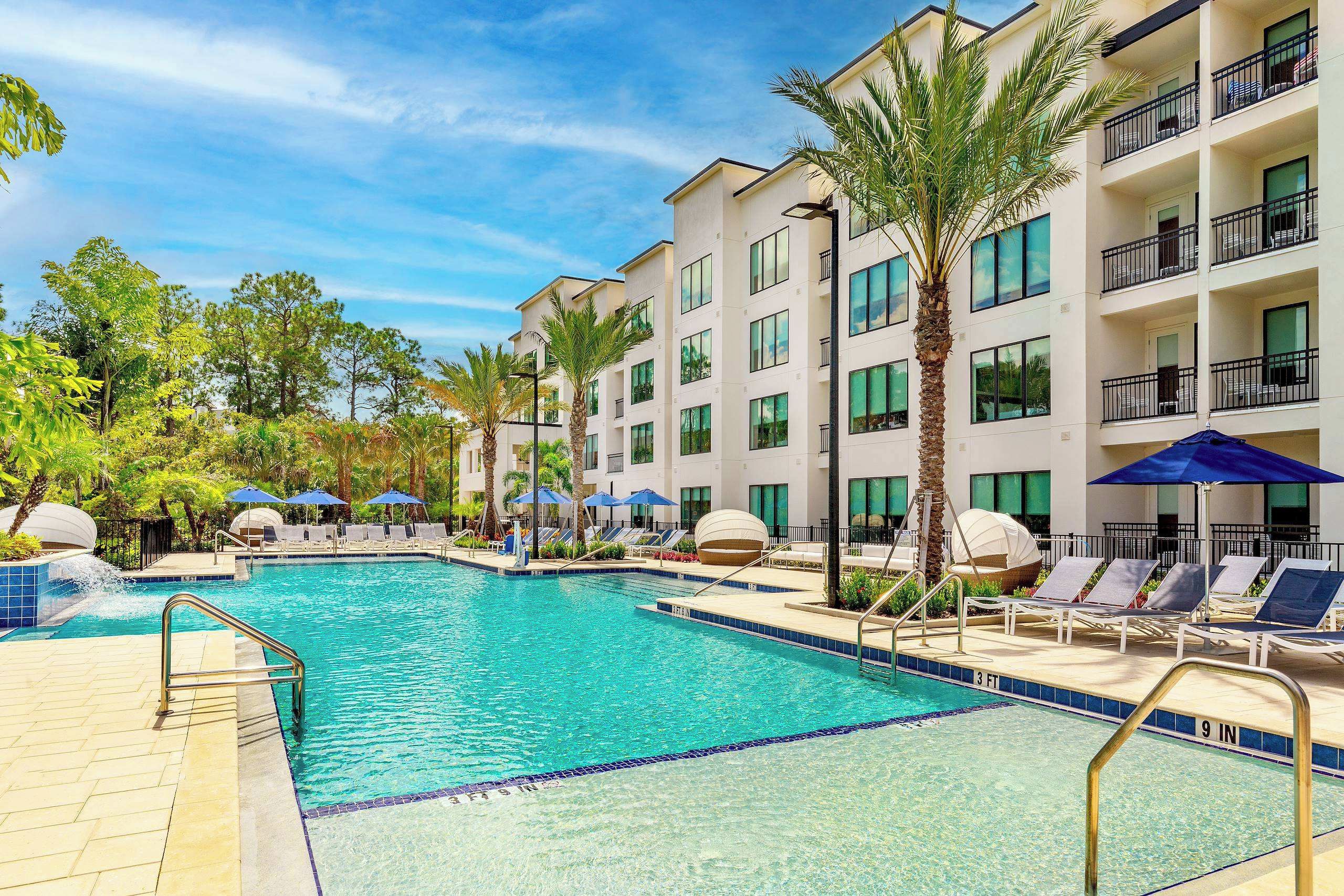 A swimming pool with lounge chairs and umbrellas, surrounded by a multi-story building with balconies.