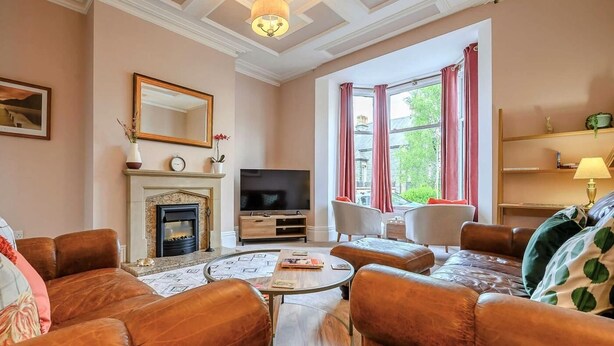 A living room with leather sofas, a fireplace, a television, and a large window with curtains. - 3 Bed in Keswick (oc-86867) (Keswick)