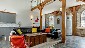 Un'ampia zona giorno con soffitto alto, travi in legno a vista, un divano in pelle nera con cuscini colorati, una poltrona rossa e un muro in pietra con una finestra.
