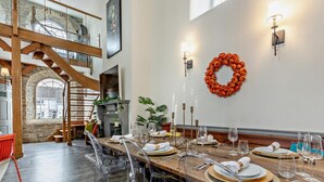 Un'area pranzo con un lungo tavolo in legno apparecchiato per sei persone, sedie trasparenti, un camino, una scala e una ghirlanda decorativa al muro.