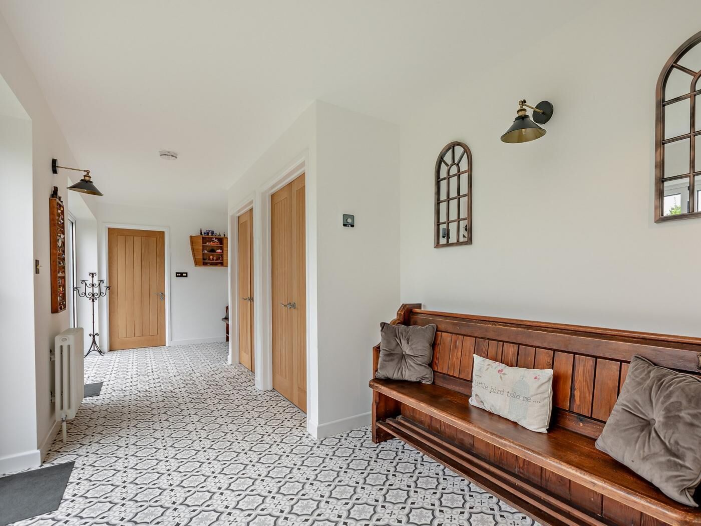 A hallway with patterned flooring, wooden doors, a bench with cushions, wall-mounted lights, and a radiator.
