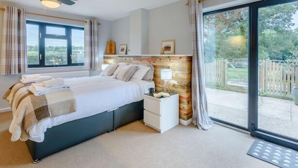 Une chambre à coucher comprenant un lit, des tables de chevet, une tête de lit en bois, un ventilateur de plafond et une grande fenêtre avec des rideaux.