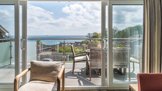 Een balkon met uitzicht op zee, houten meubilair en een glazen deur.