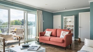 Een ruime woonkamer met een grote schuifpui naar een balkon, een rode bank, een witte fauteuil en een boekenkast.