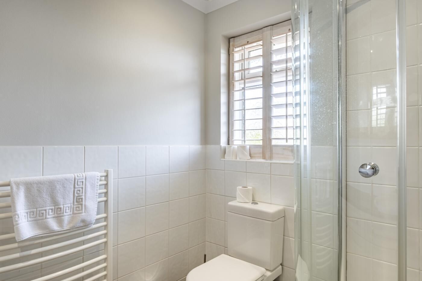 A bathroom with a toilet, a towel rack with towels, a window with blinds, and a glass shower enclosure.