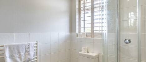 A bathroom with a toilet, a towel rack with towels, a window with blinds, and a glass shower enclosure.
