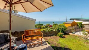 A patio with a wooden bench, a table with a plate and a bottle, and a large umbrella. - 4 Bed in Bridgend (oc-87141) (Ogmore-by-Sea)