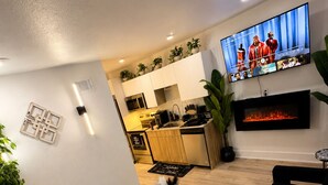 A modern living room with a black sofa, a coffee table, and a TV displaying a video call. A fireplace is visible in the background, and a kitchen area is partially seen. - Modern Luxe Escape with Spa Shower, Digital Fireplace & Prime Location (Dallas)