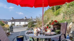 A round table with chairs, a red umbrella, and a view of a white house with chimneys. - 2 Bed in Ambleside (oc-87180) (Ambleside)