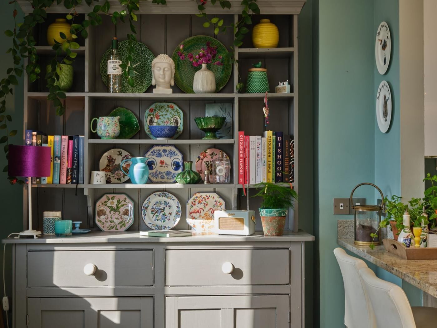 Een keuken met een wit kastje, planken met decoratieve borden, boeken en een kleine plant.