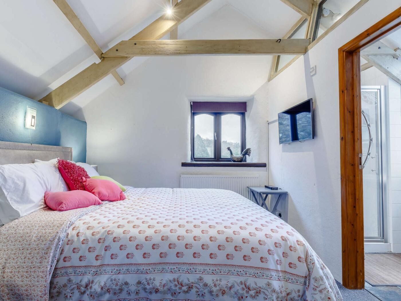A bedroom with a sloped ceiling, a bed with patterned bedding, a window with a view, and a television mounted on the wall.