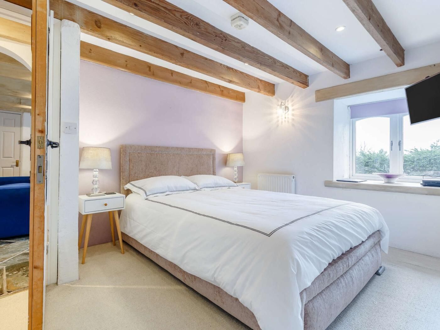 A bedroom with a large bed, wooden beams, a nightstand with a lamp, a window with a view, and a TV mounted on the wall.